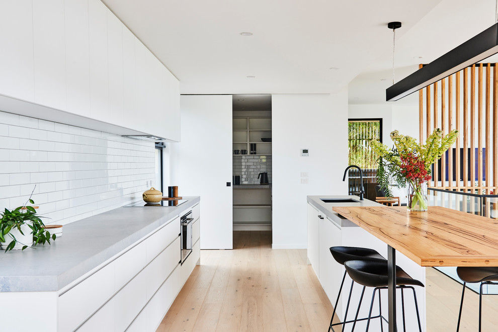 Design ideas for a mid-sized modern galley open plan kitchen in Geelong with a double-bowl sink, flat-panel cabinets, white cabinets, wood benchtops, white splashback, subway tile splashback, black appliances, light hardwood floors, with island, brown floor and grey benchtop.
