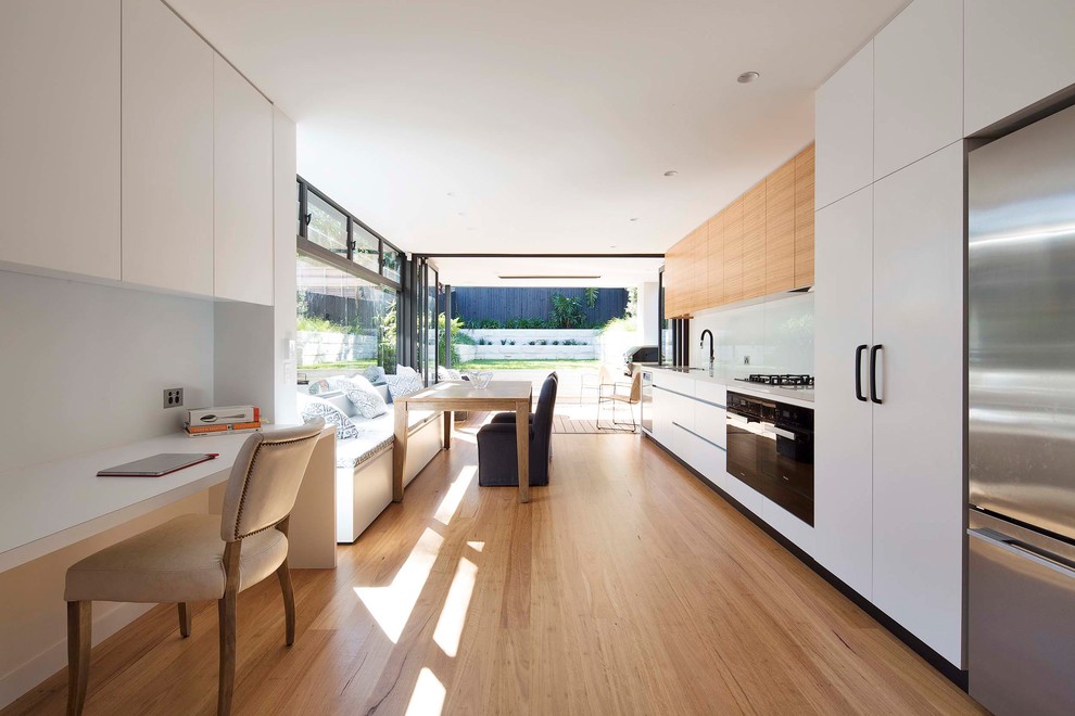 This is an example of a modern single-wall eat-in kitchen in Sydney with an undermount sink, flat-panel cabinets, quartz benchtops, white splashback, glass sheet splashback, black appliances, medium hardwood floors, no island, light wood cabinets and beige floor.