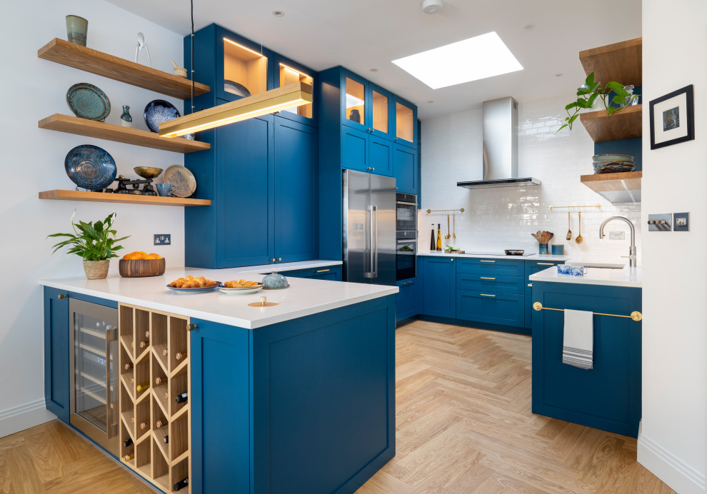 Inspiration for a large modern u-shaped open plan kitchen in London with an integrated sink, shaker cabinets, blue cabinets, solid surface benchtops, white splashback, terra-cotta splashback, stainless steel appliances, light hardwood floors, with island, brown floor and white benchtop.