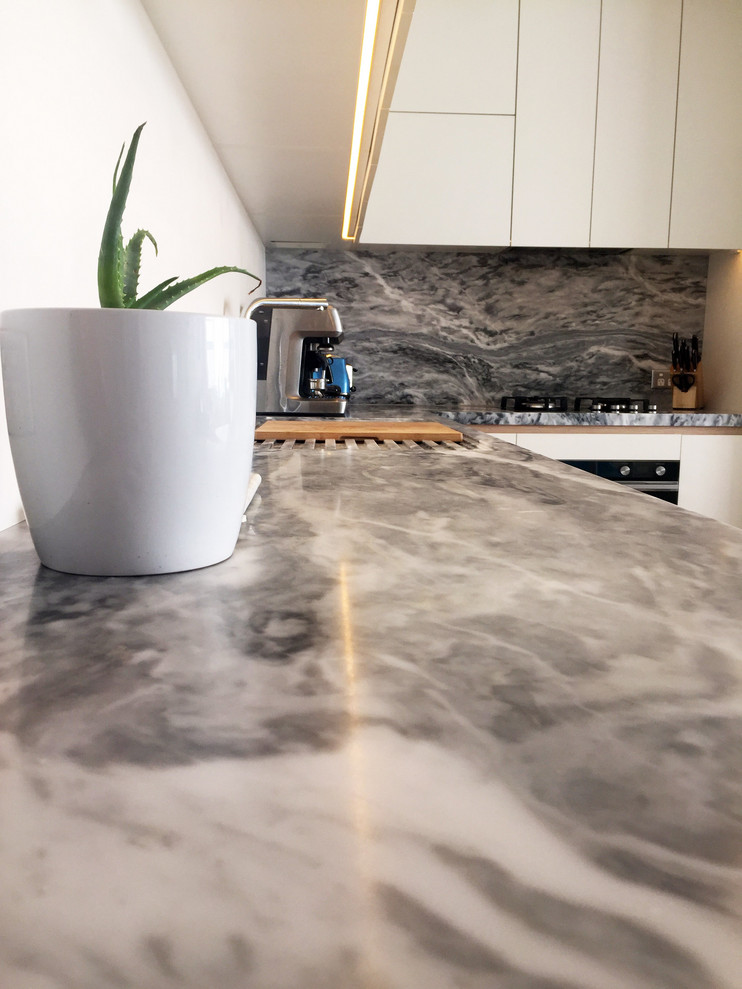 Small contemporary l-shaped eat-in kitchen in Sydney with a double-bowl sink, flat-panel cabinets, white cabinets, marble benchtops, white splashback, marble splashback, black appliances, medium hardwood floors, no island, brown floor and grey benchtop.