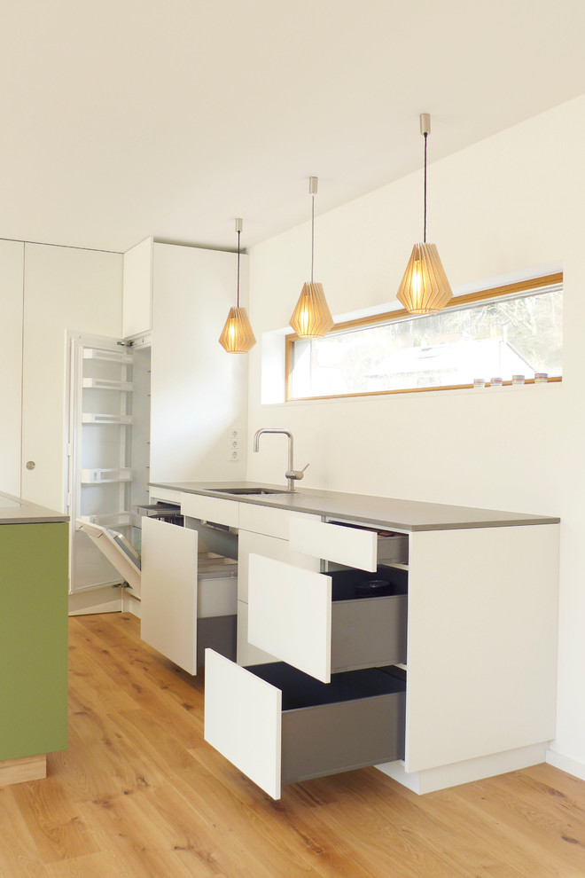 Inspiration for a small scandinavian galley eat-in kitchen in Frankfurt with an undermount sink, flat-panel cabinets, green cabinets, quartz benchtops, white splashback, timber splashback, black appliances, medium hardwood floors, with island, brown floor and grey benchtop.