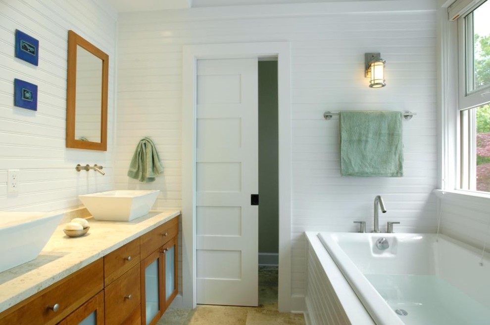 Mid-sized beach style master bathroom in New York with glass-front cabinets, medium wood cabinets, limestone benchtops, beige tile, stone slab, a drop-in tub, a one-piece toilet, a vessel sink, white walls, travertine floors, a double shower and beige floor.
