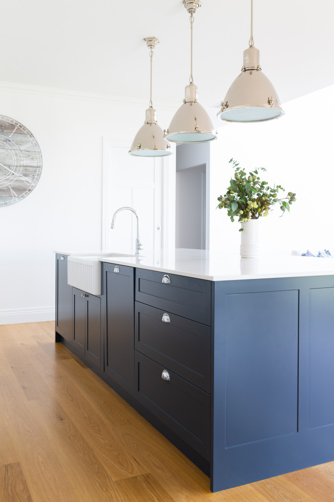 Inspiration for a traditional galley kitchen in Canberra - Queanbeyan with a farmhouse sink, shaker cabinets, blue cabinets, quartz benchtops, white splashback, subway tile splashback, stainless steel appliances, light hardwood floors, with island and white benchtop.