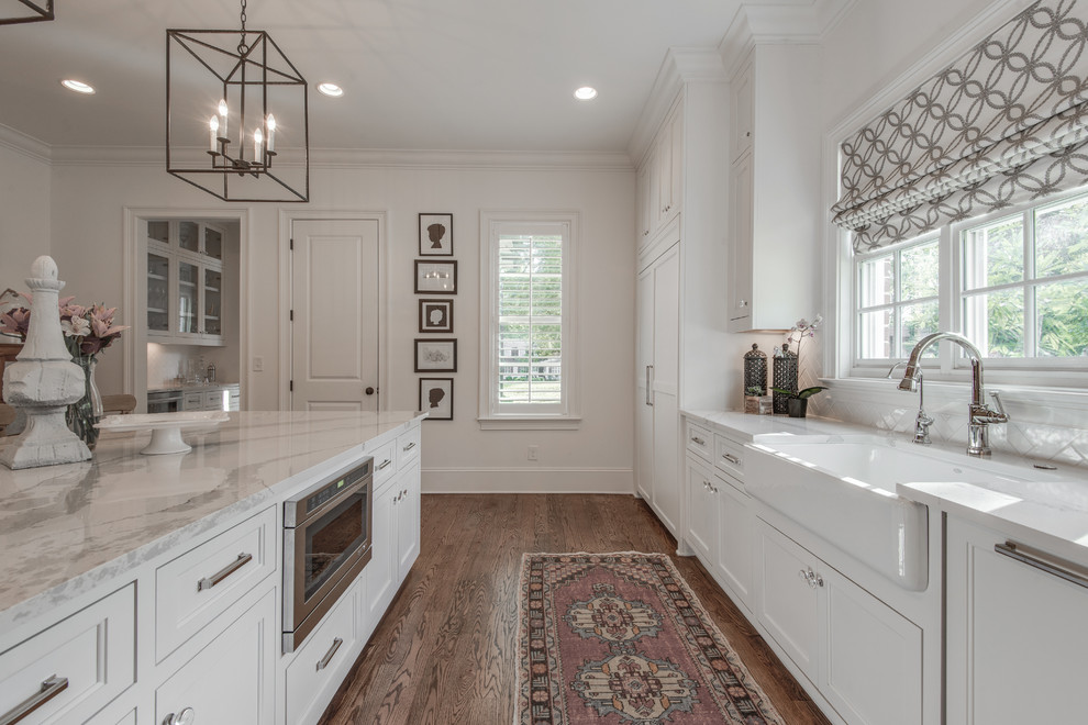 Inspiration for a large transitional l-shaped kitchen in Nashville with a farmhouse sink, recessed-panel cabinets, white cabinets, quartz benchtops, white splashback, porcelain splashback, panelled appliances, medium hardwood floors, with island and brown floor.