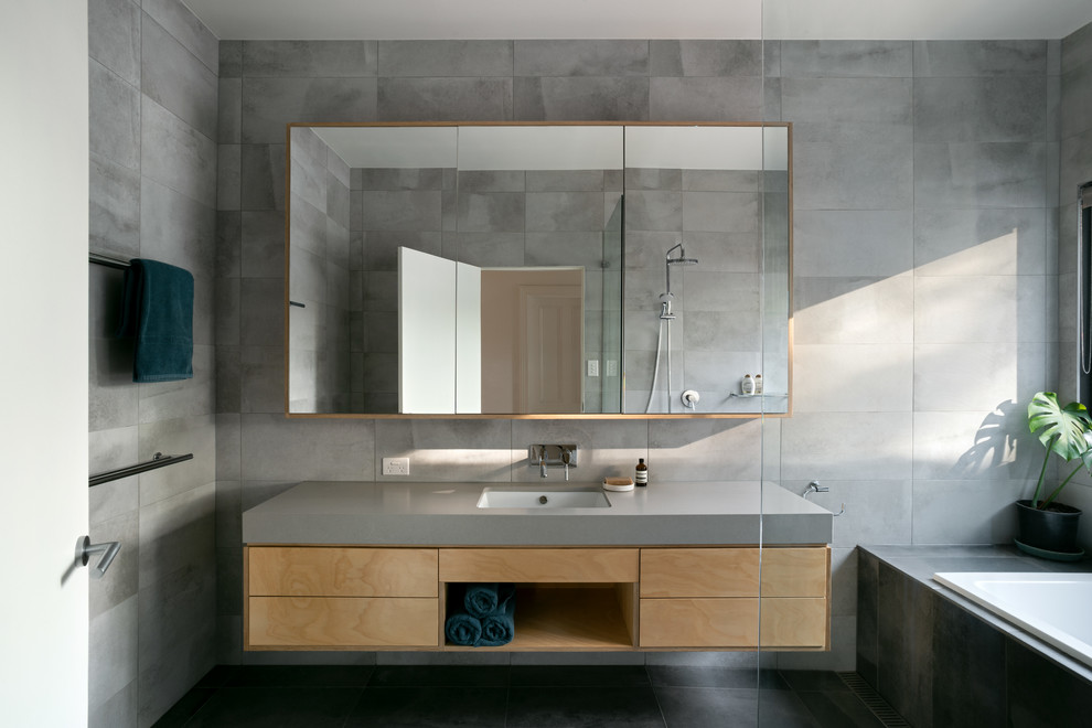 This is an example of a contemporary bathroom in Melbourne with flat-panel cabinets, medium wood cabinets, a drop-in tub, gray tile, grey walls, an undermount sink, black floor and grey benchtops.