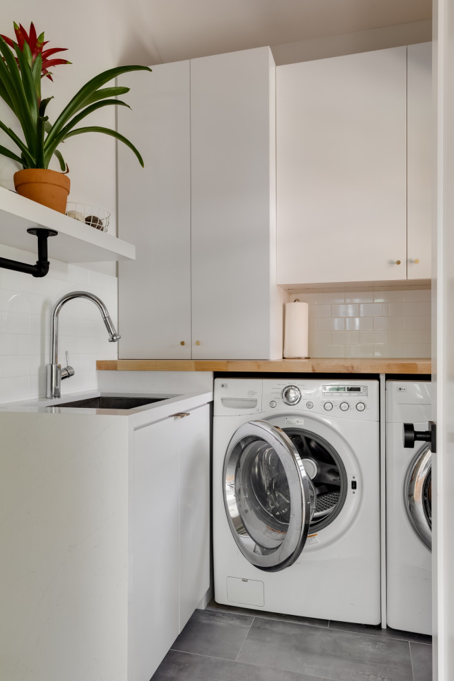 Inspiration for a small contemporary l-shaped dedicated laundry room in Los Angeles with an undermount sink, flat-panel cabinets, white cabinets, wood benchtops, white splashback, subway tile splashback, white walls, ceramic floors, a side-by-side washer and dryer, grey floor, vaulted and brown benchtop.