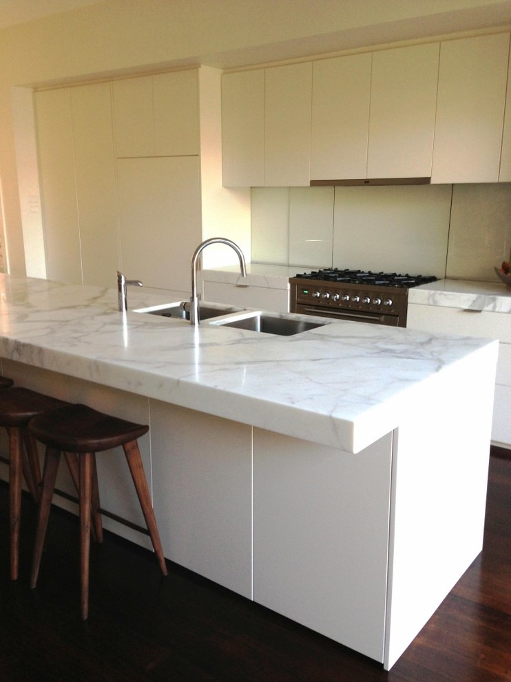 Photo of a contemporary galley kitchen in Melbourne with flat-panel cabinets, white cabinets, marble benchtops, white splashback, glass sheet splashback, stainless steel appliances and with island.