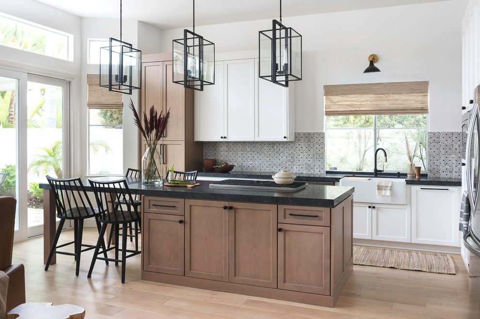 Photo of a mid-sized transitional l-shaped eat-in kitchen in San Diego with a farmhouse sink, shaker cabinets, light wood cabinets, granite benchtops, beige splashback, terra-cotta splashback, stainless steel appliances, light hardwood floors, with island and black benchtop.