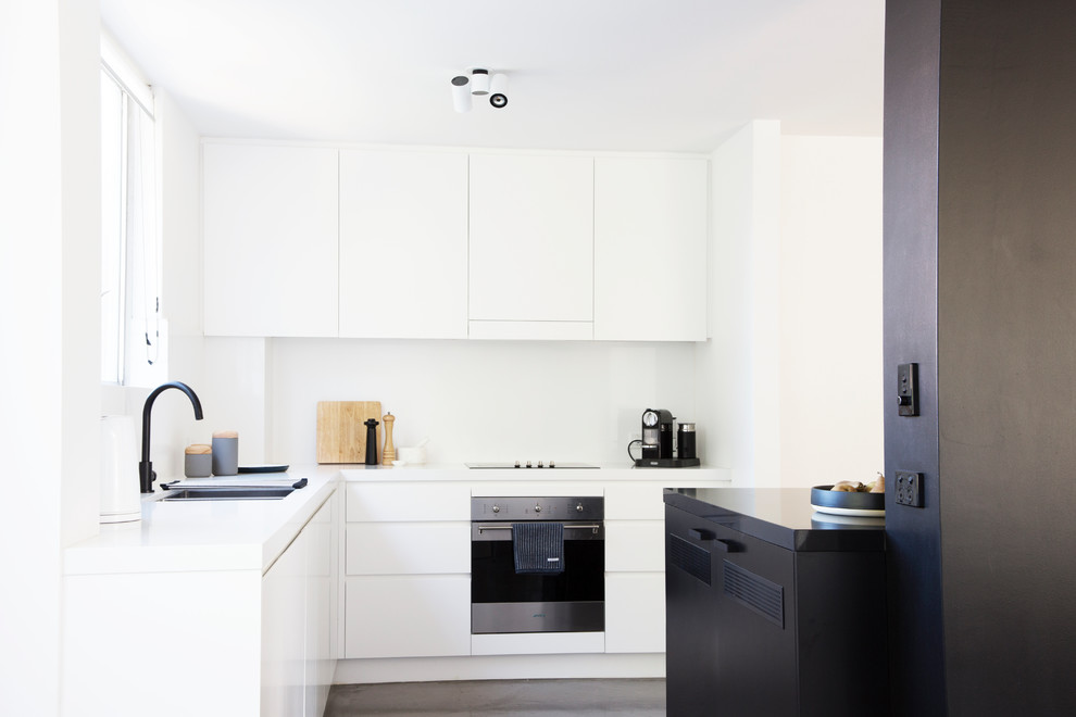 Photo of a contemporary l-shaped kitchen in Sydney with a drop-in sink, flat-panel cabinets, white cabinets, white splashback, glass sheet splashback, black appliances, concrete floors, with island, grey floor and white benchtop.