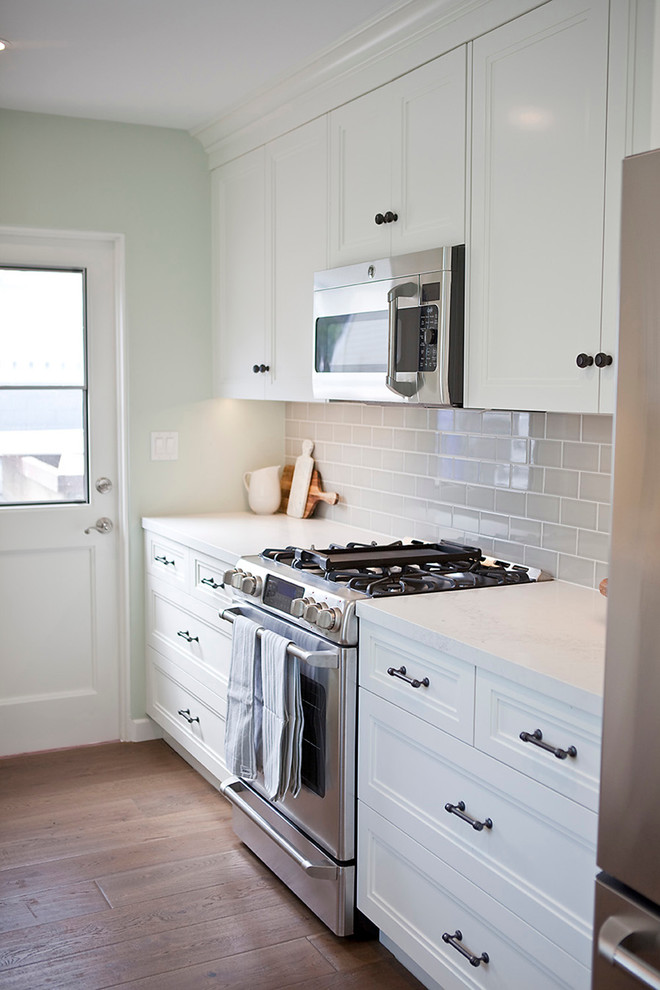 Photo of a mid-sized beach style galley separate kitchen in San Diego with a farmhouse sink, beaded inset cabinets, white cabinets, quartz benchtops, grey splashback, ceramic splashback, stainless steel appliances, medium hardwood floors and no island.