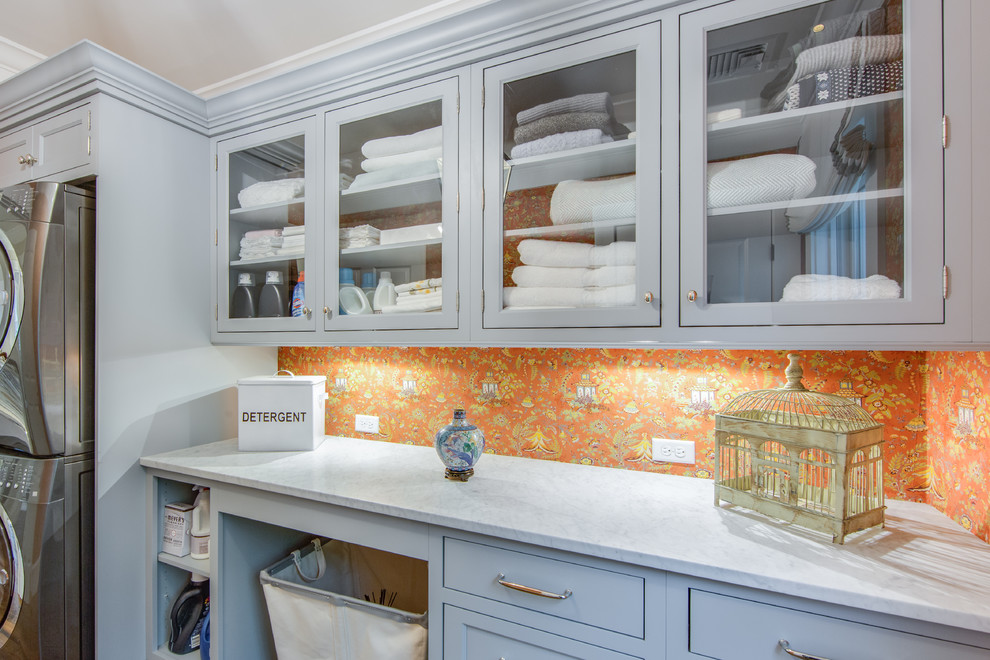 Large transitional laundry room in Boston with a stacked washer and dryer, glass-front cabinets, marble benchtops and grey cabinets.