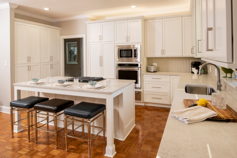 Large transitional l-shaped eat-in kitchen in Raleigh with an undermount sink, shaker cabinets, white cabinets, quartz benchtops, grey splashback, ceramic splashback, stainless steel appliances, with island, brown floor and grey benchtop.