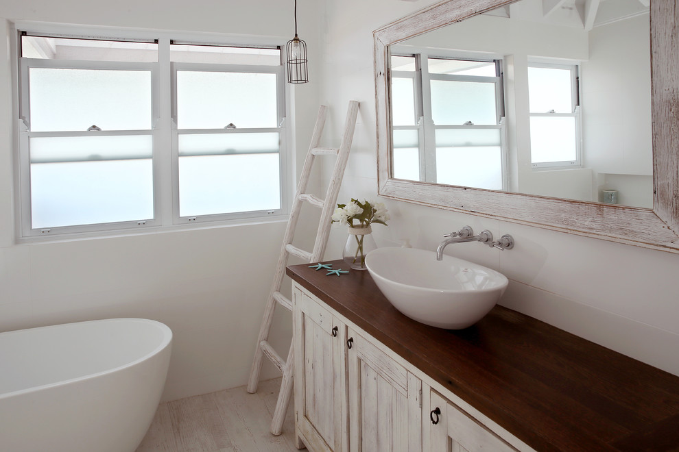 This is an example of a large beach style 3/4 bathroom in Sydney with furniture-like cabinets, distressed cabinets, a freestanding tub, white tile, white walls, light hardwood floors, a trough sink, wood benchtops and brown benchtops.