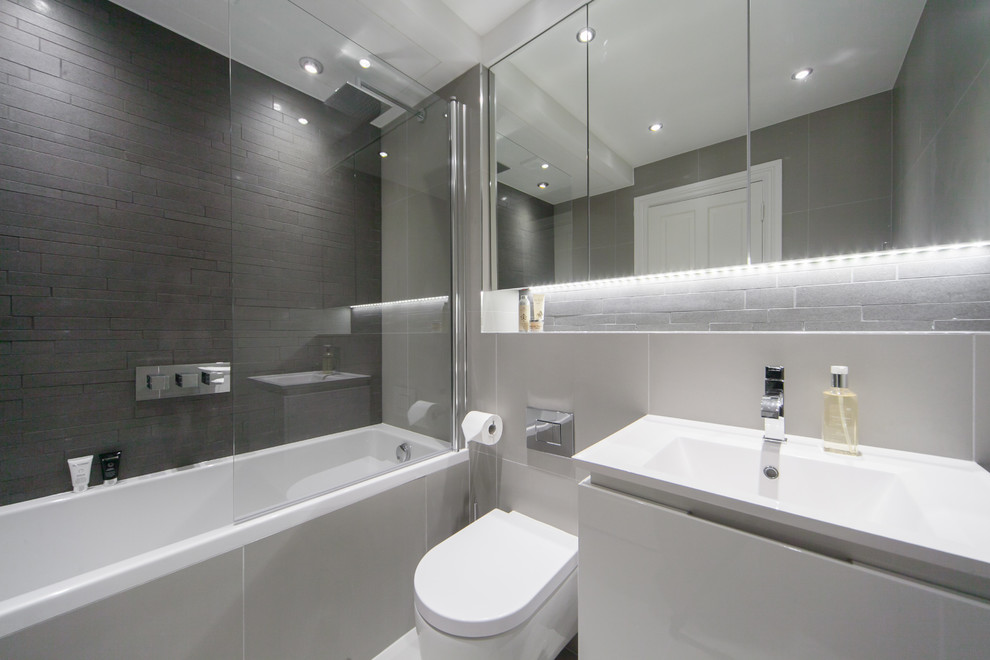 Mid-sized contemporary master bathroom in London with a wall-mount sink, furniture-like cabinets, grey cabinets, an alcove tub, an alcove shower, a wall-mount toilet, gray tile, ceramic tile, grey walls and ceramic floors.