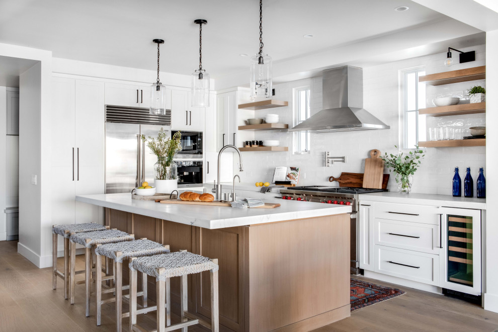 Example of a transitional l-shaped light wood floor and beige floor kitchen design in Orange County with a farmhouse sink, shaker cabinets, white cabinets, white backsplash, stainless steel appliances, an island and white countertops