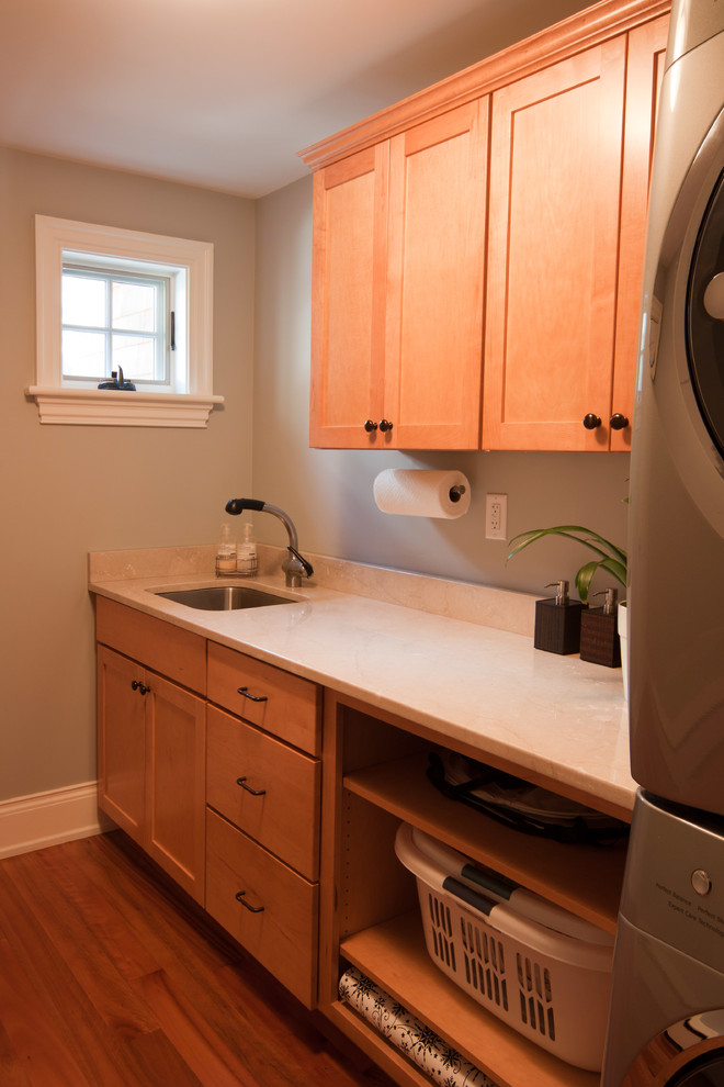 Photo of a mid-sized traditional single-wall dedicated laundry room in Boston with an undermount sink, shaker cabinets, solid surface benchtops, beige walls, light hardwood floors, a stacked washer and dryer and medium wood cabinets.