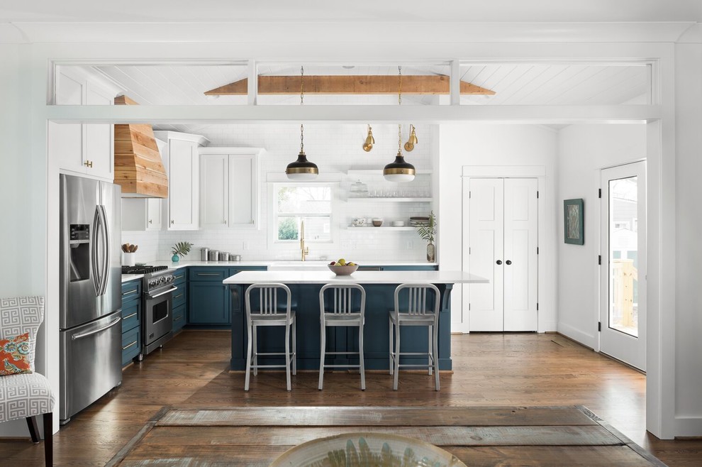 This is an example of a country l-shaped open plan kitchen in New Orleans with a farmhouse sink, shaker cabinets, blue cabinets, white splashback, subway tile splashback, stainless steel appliances, medium hardwood floors, with island, brown floor, white benchtop and marble benchtops.