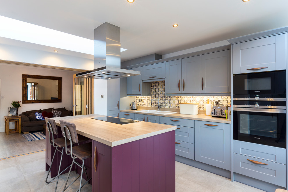 Photo of a transitional galley kitchen in London with a drop-in sink, shaker cabinets, blue cabinets, wood benchtops, multi-coloured splashback, ceramic splashback, black appliances and with island.