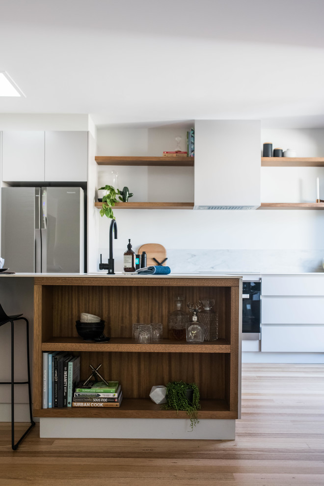 This is an example of a mid-sized contemporary galley open plan kitchen in Melbourne with a single-bowl sink, shaker cabinets, quartz benchtops, white splashback, stone slab splashback, stainless steel appliances, light hardwood floors, with island, brown floor and white benchtop.