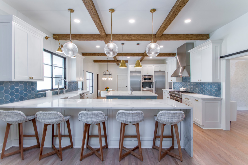 This is an example of a beach style kitchen in Little Rock with a farmhouse sink, shaker cabinets, white cabinets, quartzite benchtops, blue splashback, ceramic splashback, stainless steel appliances, white benchtop, light hardwood floors and a peninsula.