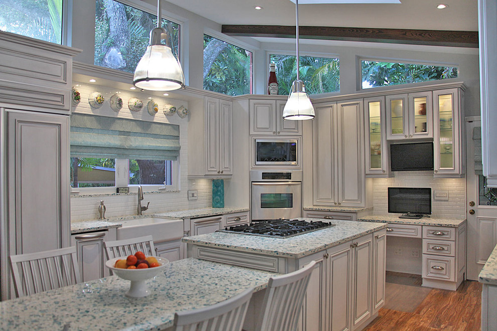 Photo of a beach style u-shaped eat-in kitchen in Miami with a farmhouse sink, white cabinets, recycled glass benchtops, white splashback, subway tile splashback and stainless steel appliances.