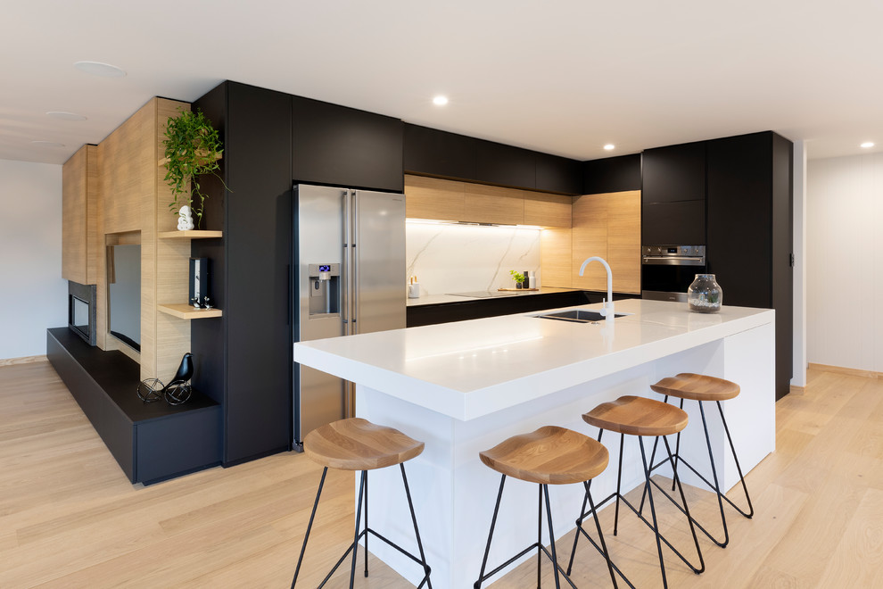 This is an example of a large contemporary l-shaped eat-in kitchen in Other with an undermount sink, flat-panel cabinets, black cabinets, solid surface benchtops, white splashback, ceramic splashback, stainless steel appliances, light hardwood floors, with island, white benchtop and beige floor.