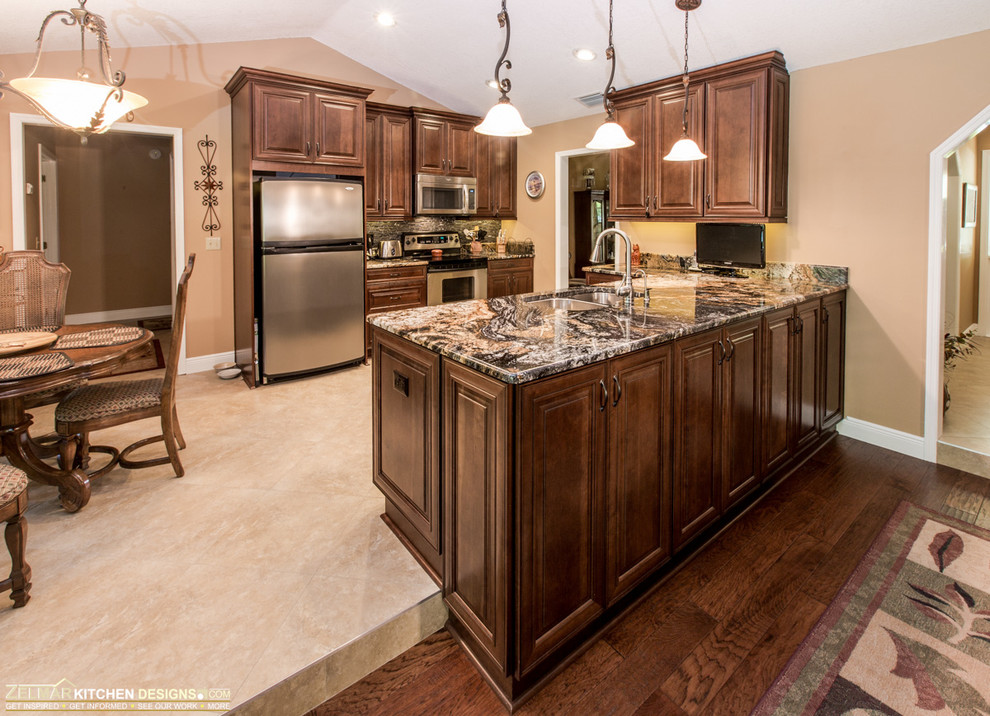 Photo of a mid-sized transitional u-shaped eat-in kitchen in Orlando with an undermount sink, raised-panel cabinets, medium wood cabinets, granite benchtops, porcelain floors, grey splashback, glass sheet splashback, stainless steel appliances and a peninsula.