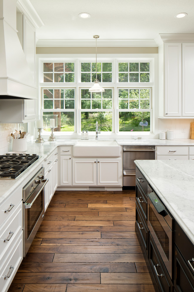 This is an example of a traditional l-shaped kitchen in Other with a farmhouse sink, recessed-panel cabinets, white cabinets, white splashback, subway tile splashback, stainless steel appliances, dark hardwood floors, with island and brown floor.