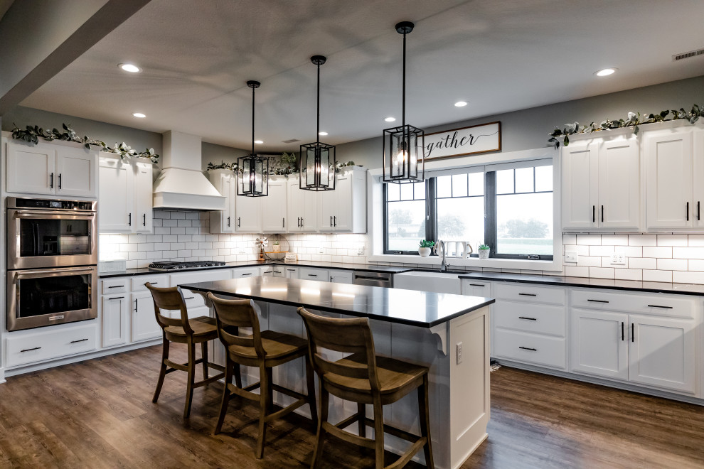Transitional u-shaped medium tone wood floor and brown floor enclosed kitchen photo with a farmhouse sink, shaker cabinets, white cabinets, white backsplash, subway tile backsplash, stainless steel appliances, an island and black countertops