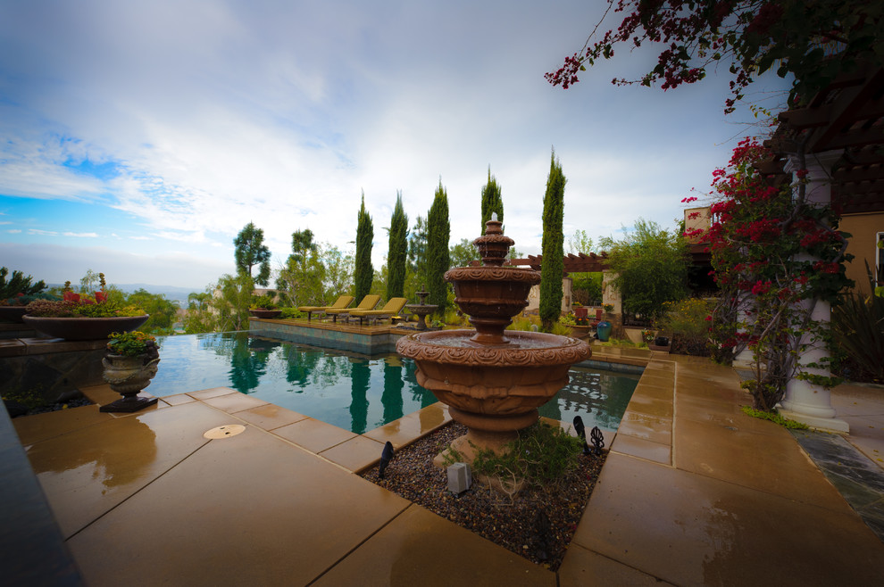 Photo of a large mediterranean backyard custom-shaped infinity pool in Orange County with a water feature and concrete slab.