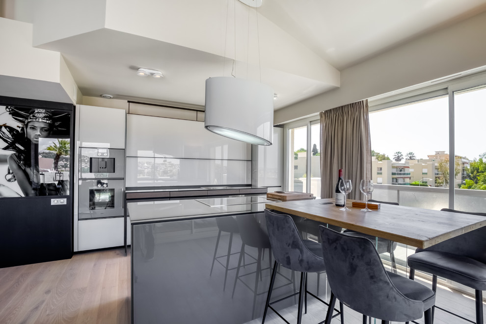 Inspiration for a large contemporary galley eat-in kitchen in Nice with an undermount sink, glass benchtops, white splashback, black appliances, light hardwood floors, grey benchtop and with island.