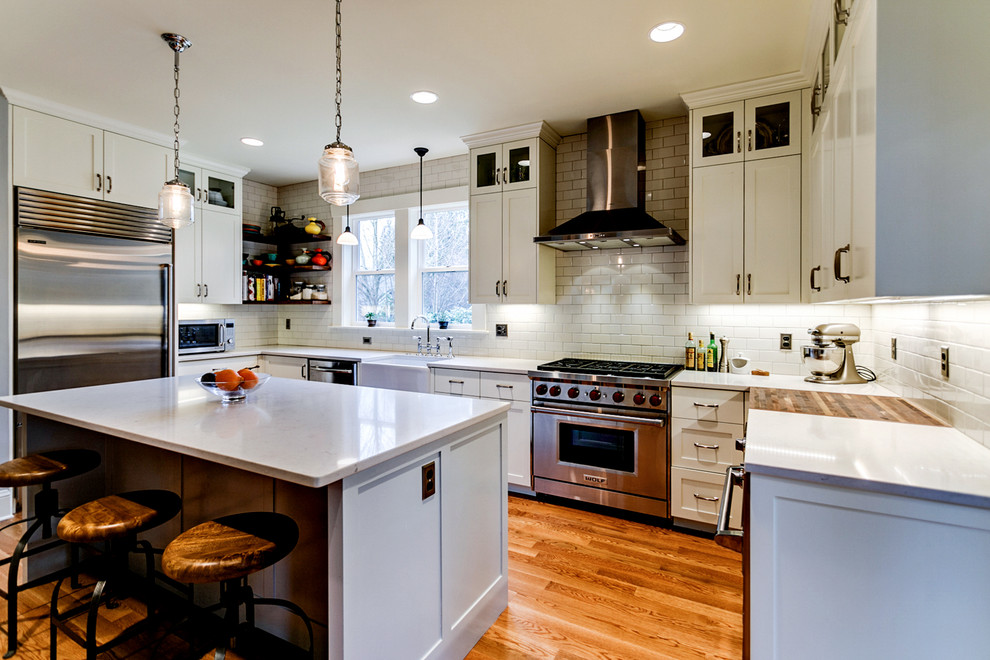 This is an example of a mid-sized industrial l-shaped eat-in kitchen in Portland with a farmhouse sink, shaker cabinets, white cabinets, quartz benchtops, white splashback, ceramic splashback, stainless steel appliances, medium hardwood floors, with island and brown floor.