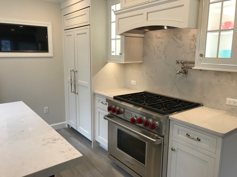 Mid-sized transitional u-shaped light wood floor and brown floor open concept kitchen photo with a farmhouse sink, recessed-panel cabinets, white cabinets, marble countertops, white backsplash, marble backsplash, stainless steel appliances and an island