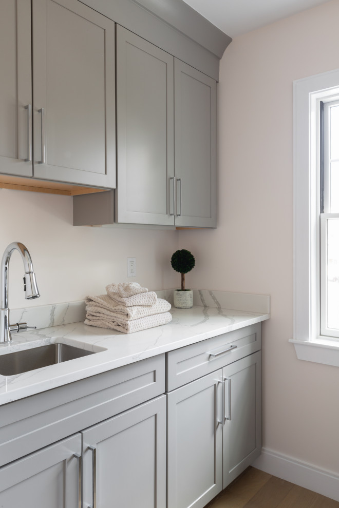 This is an example of a medium sized traditional galley separated utility room in Boston with a submerged sink, shaker cabinets, grey cabinets, engineered stone countertops, white splashback, granite splashback, beige walls, light hardwood flooring, a side by side washer and dryer, beige floors and white worktops.