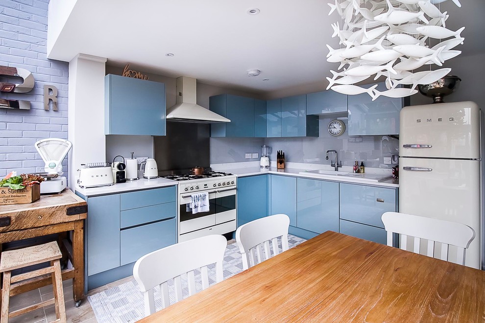 This is an example of a mid-sized eclectic l-shaped open plan kitchen in London with an integrated sink, flat-panel cabinets, blue cabinets, solid surface benchtops, grey splashback, glass sheet splashback, white appliances, travertine floors, beige floor and white benchtop.