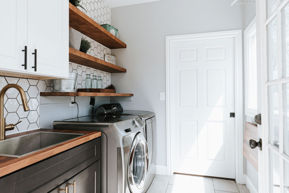 This is an example of a small contemporary single-wall dedicated laundry room in Denver with a drop-in sink, shaker cabinets, white cabinets, wood benchtops, white splashback, subway tile splashback, grey walls, porcelain floors, a side-by-side washer and dryer, grey floor and brown benchtop.