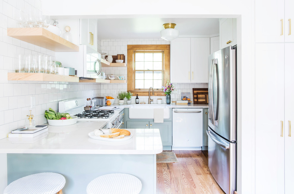 Photo of a small transitional u-shaped eat-in kitchen in Other with a farmhouse sink, shaker cabinets, blue cabinets, quartz benchtops, white splashback, porcelain splashback, white appliances, medium hardwood floors, a peninsula and brown floor.