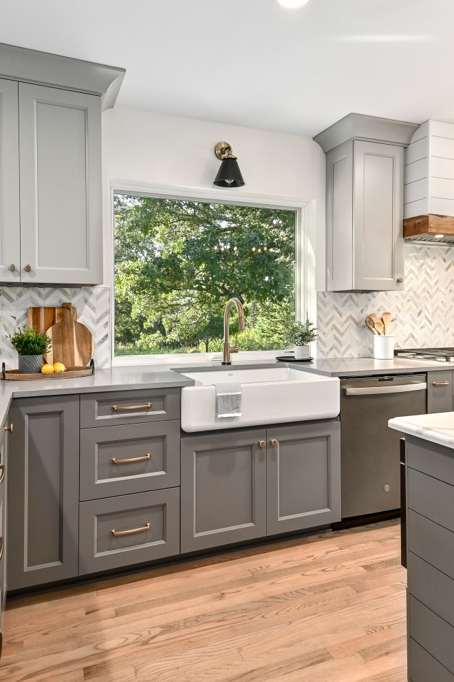 Design ideas for an expansive country l-shaped eat-in kitchen in Milwaukee with a farmhouse sink, shaker cabinets, grey cabinets, quartz benchtops, white splashback, marble splashback, stainless steel appliances, medium hardwood floors, with island, brown floor and grey benchtop.