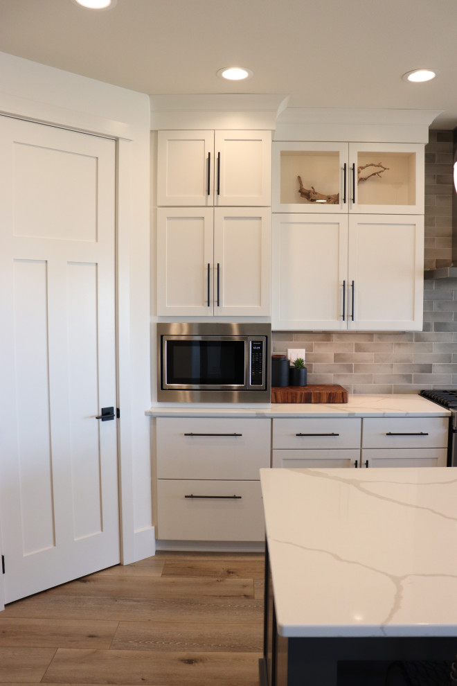 Large transitional l-shaped medium tone wood floor and brown floor open concept kitchen photo with a single-bowl sink, shaker cabinets, white cabinets, quartz countertops, gray backsplash, subway tile backsplash, stainless steel appliances, an island and white countertops