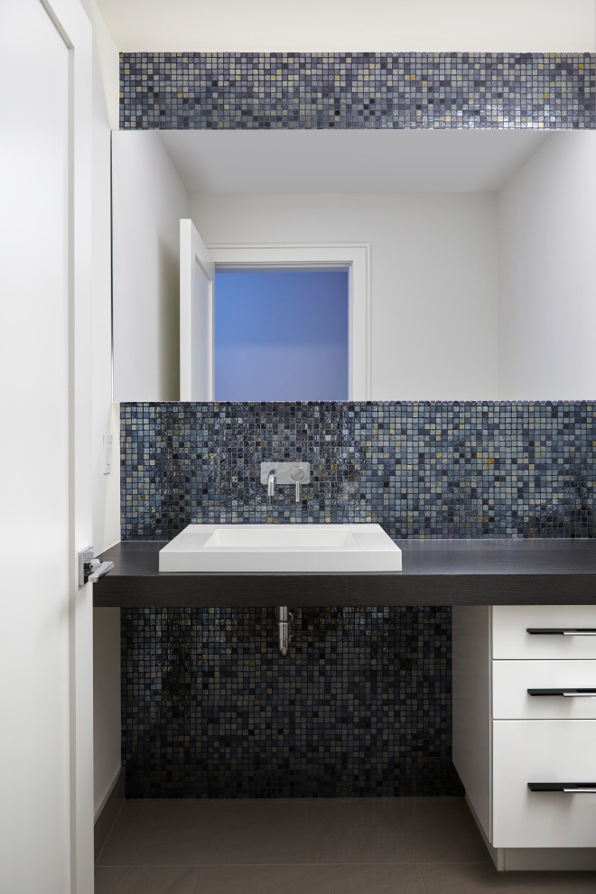 This is an example of a mid-sized contemporary powder room in Toronto with flat-panel cabinets, white cabinets, a wall-mount toilet, black tile, mirror tile, white walls, porcelain floors, a vessel sink, laminate benchtops, grey floor and grey benchtops.