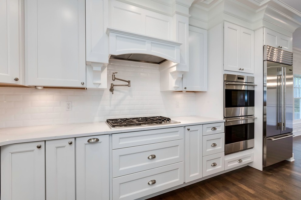 Photo of a large transitional l-shaped open plan kitchen in New York with a farmhouse sink, shaker cabinets, white cabinets, quartz benchtops, white splashback, subway tile splashback, stainless steel appliances, medium hardwood floors, with island and brown floor.