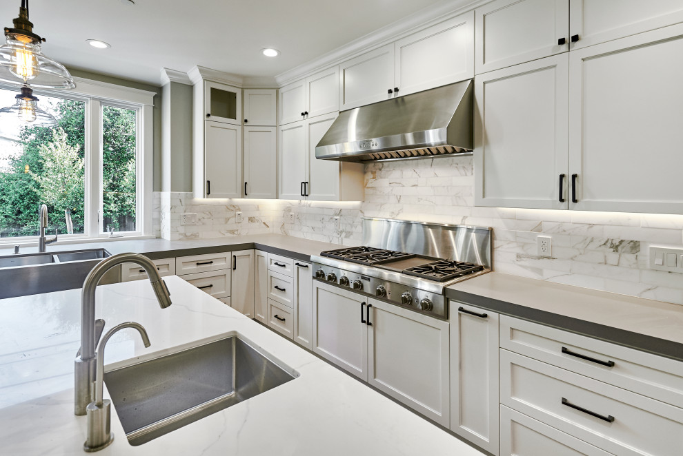 Mid-sized arts and crafts l-shaped open plan kitchen in San Francisco with a farmhouse sink, shaker cabinets, white cabinets, quartz benchtops, grey splashback, subway tile splashback, stainless steel appliances, medium hardwood floors, with island, brown floor and grey benchtop.