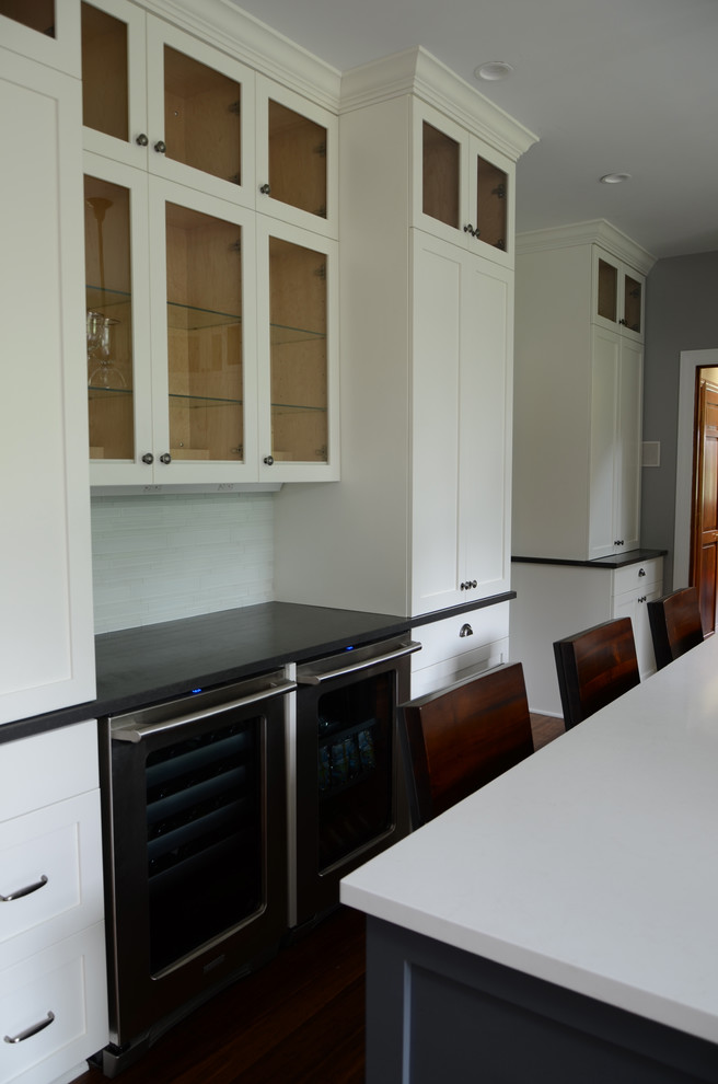 Mid-sized transitional l-shaped dark wood floor eat-in kitchen photo with a single-bowl sink, shaker cabinets, white cabinets, solid surface countertops, white backsplash, glass tile backsplash, stainless steel appliances and an island