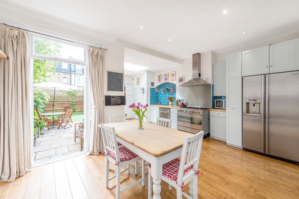 This is an example of a traditional eat-in kitchen in London with shaker cabinets, white cabinets, wood benchtops, blue splashback, subway tile splashback, stainless steel appliances and medium hardwood floors.