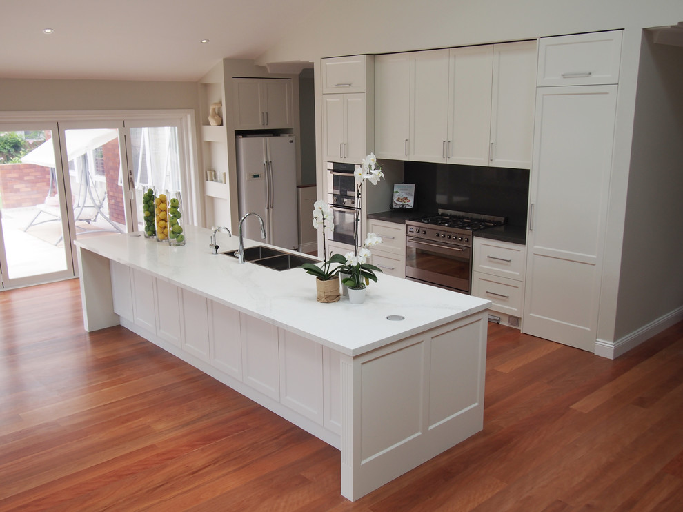 Large transitional galley kitchen pantry in Sydney with a farmhouse sink, shaker cabinets, white cabinets, quartz benchtops, grey splashback, stone slab splashback, stainless steel appliances, medium hardwood floors and with island.
