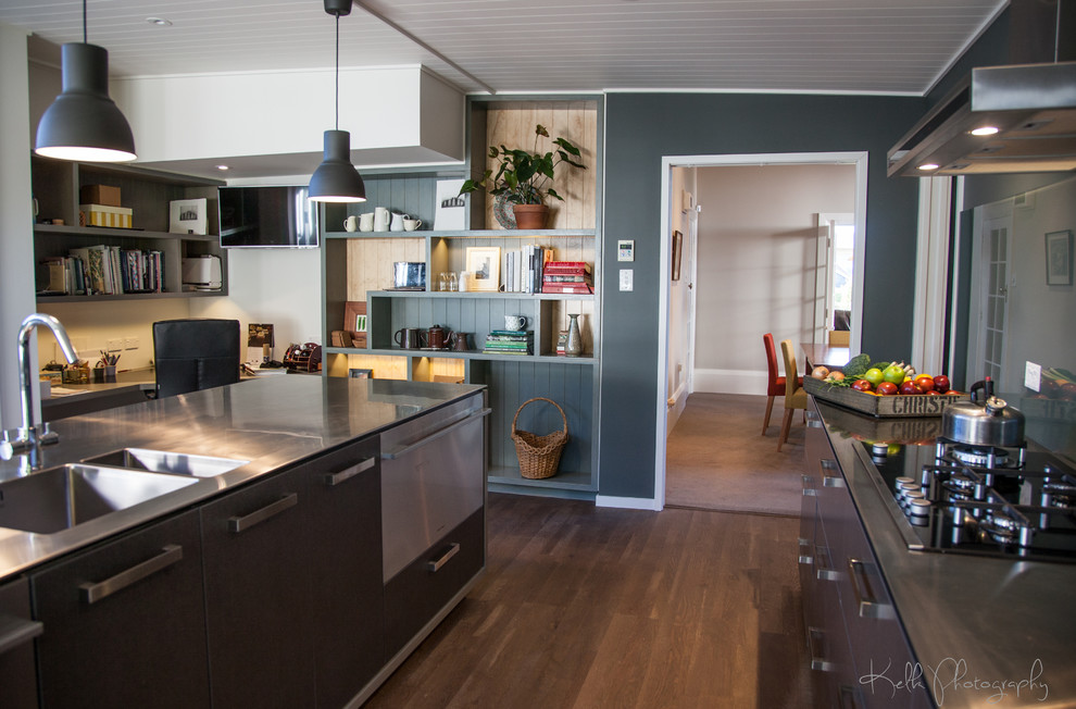 This is an example of a mid-sized industrial galley open plan kitchen in Dunedin with stainless steel benchtops, a double-bowl sink, flat-panel cabinets, grey cabinets, green splashback, glass sheet splashback, stainless steel appliances, laminate floors, with island and brown floor.
