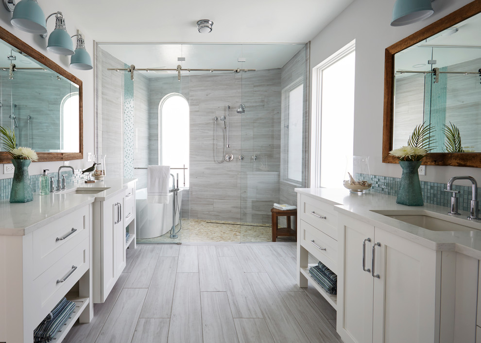 This is an example of a large beach style master wet room bathroom in Other with shaker cabinets, white cabinets, a freestanding tub, a two-piece toilet, gray tile, wood-look tile, grey walls, wood-look tile, an undermount sink, engineered quartz benchtops, grey floor, a sliding shower screen, grey benchtops, an enclosed toilet, a double vanity and a freestanding vanity.