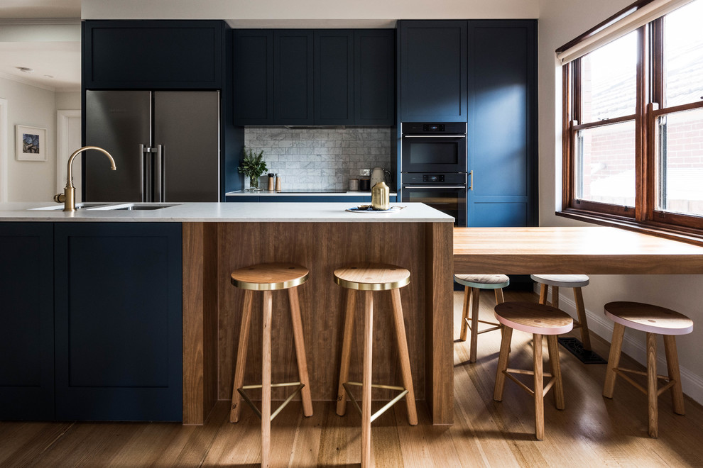 This is an example of a transitional galley kitchen in Melbourne with an undermount sink, shaker cabinets, blue cabinets, grey splashback, marble splashback, black appliances, medium hardwood floors, with island, brown floor and grey benchtop.