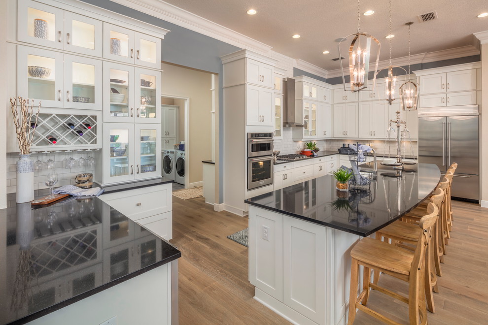 Large country u-shaped separate kitchen in Orlando with shaker cabinets, white cabinets, white splashback, subway tile splashback, stainless steel appliances, medium hardwood floors, with island, a farmhouse sink, quartz benchtops, brown floor and black benchtop.