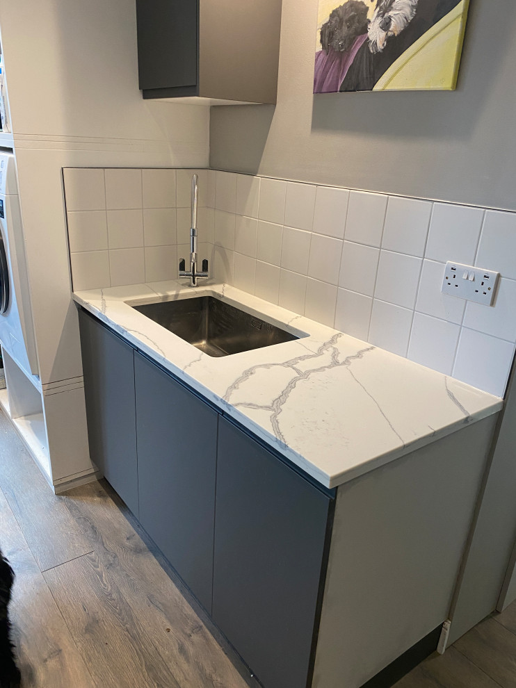 Design ideas for a large contemporary galley utility room in London with a built-in sink, flat-panel cabinets, grey cabinets, quartz worktops, white splashback, porcelain splashback, beige walls, laminate floors, a side by side washer and dryer, grey floors and multicoloured worktops.